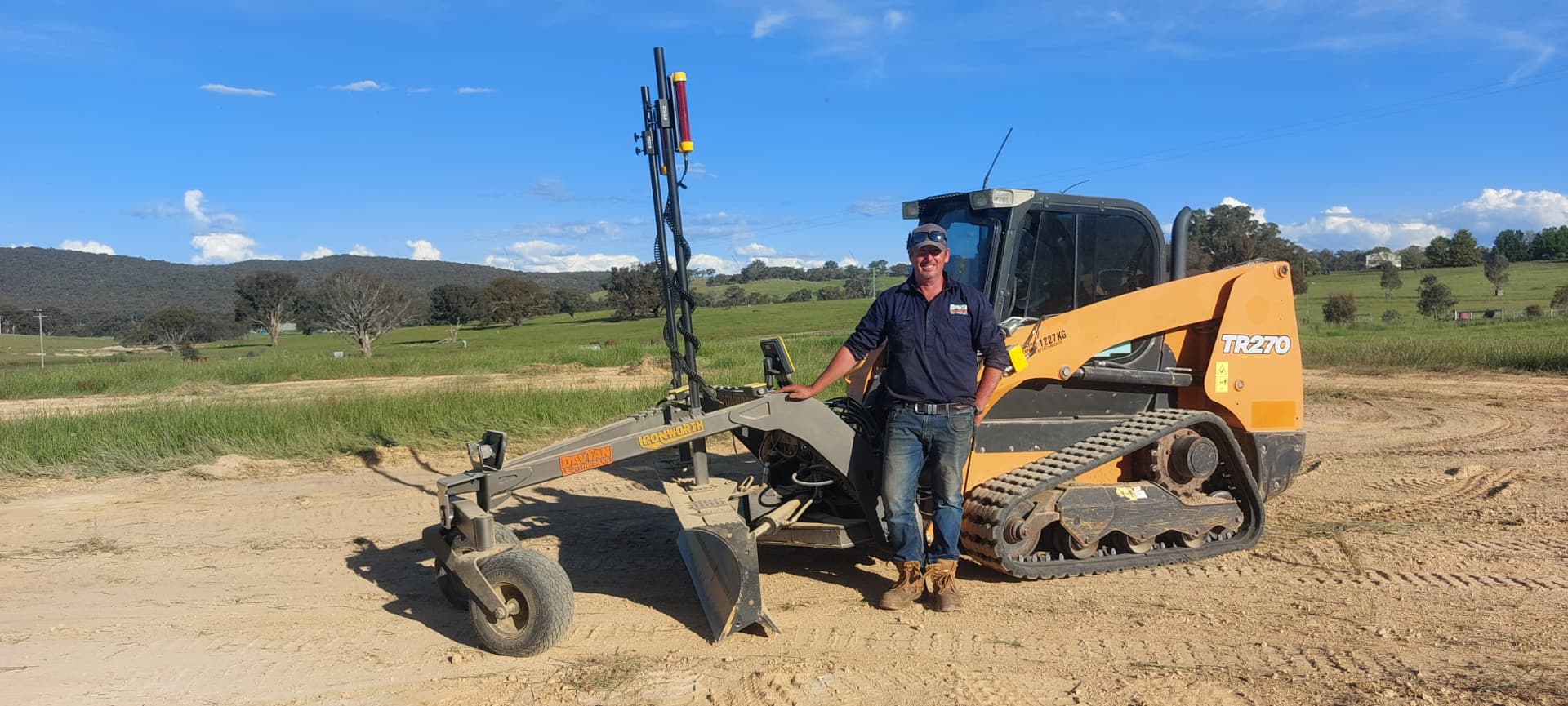 Dave Hall standing beside his Ironworth grader blade and laser mast fitted to a compact track loader on a Canberra property